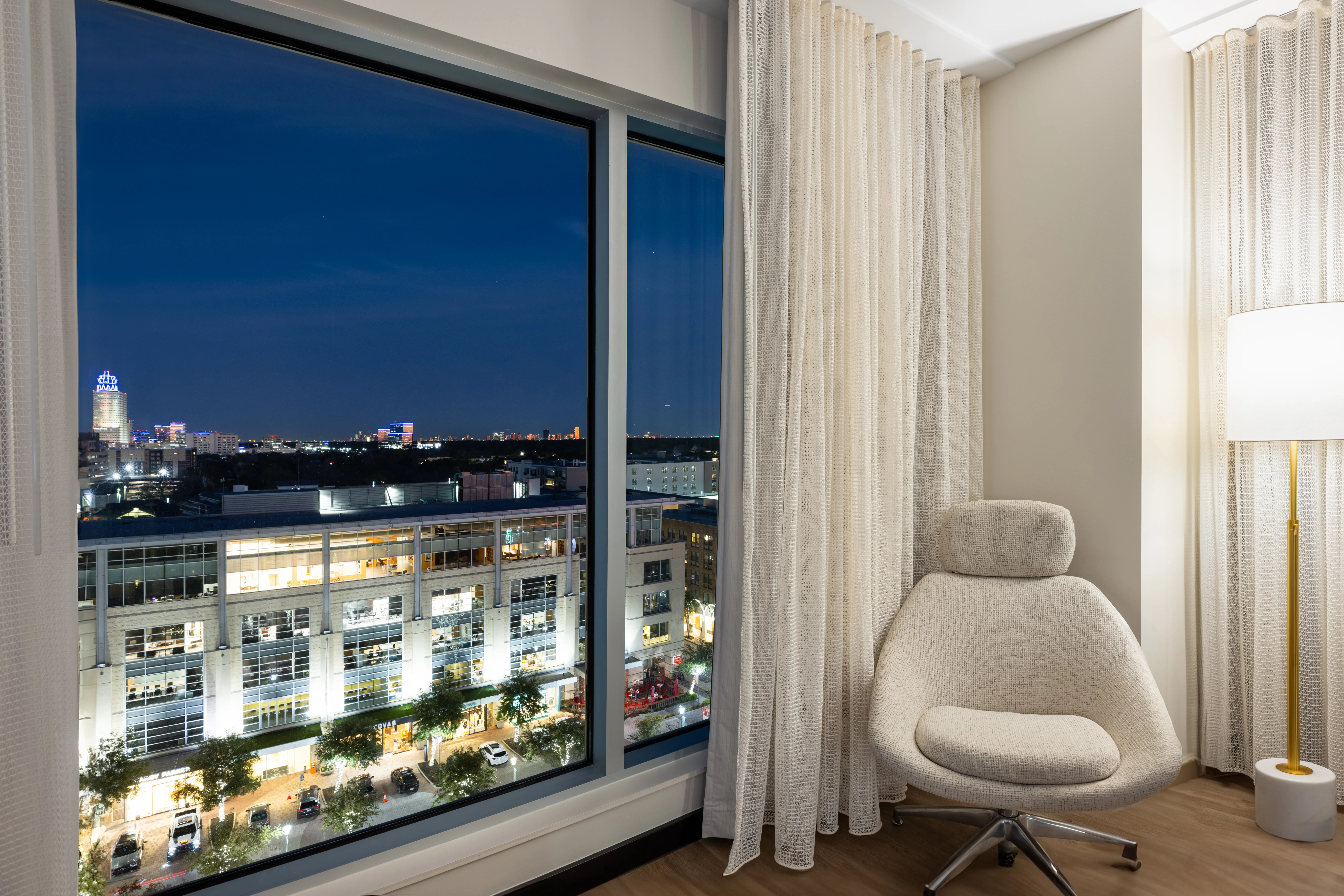 Hotel room corner with a white chair, lamp and large window.